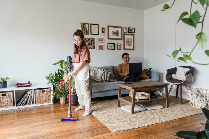 Woman cleaning living room with vacuum, man on couch with laptop, enjoying improved life quality.