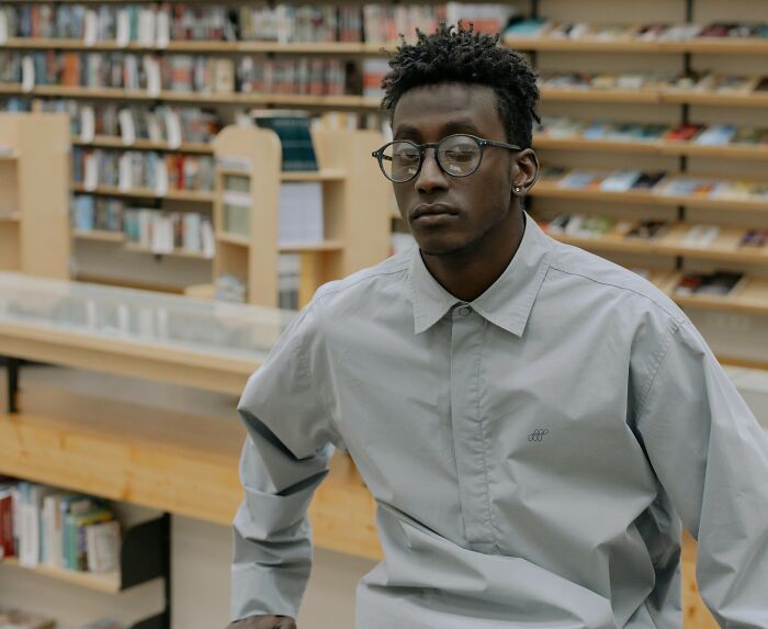 Young man in a library, wearing glasses and a light gray shirt, sitting with a thoughtful expression.