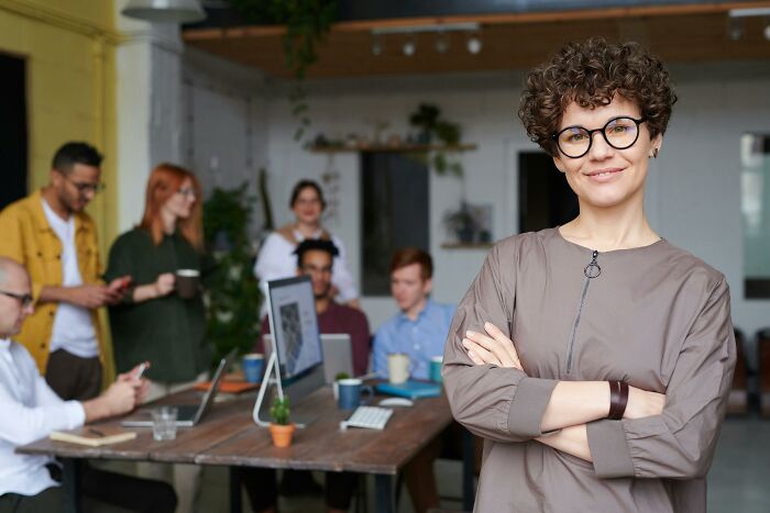 Office setting with a smiling woman in focus, colleagues discussing adult realizations near a table with laptops.