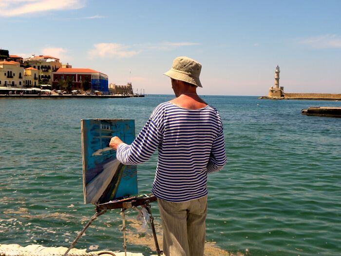 Man painting seascape at harbor, wearing striped shirt and hat, illustrating judgment-free creativity.