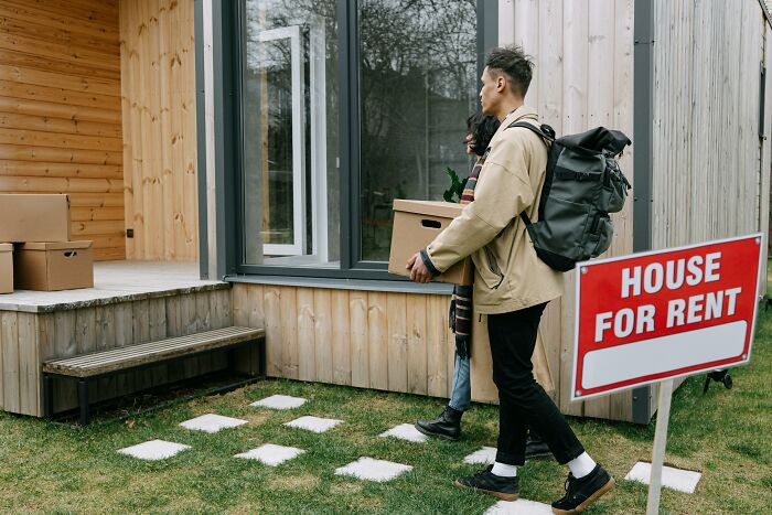 Two people carrying boxes in front of a house with a "For Rent" sign, highlighting financial stereotypes in housing.