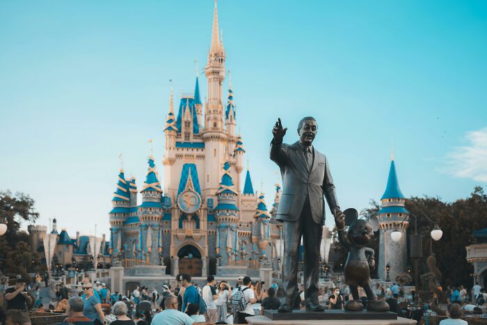 Statue of Walt Disney and Mickey Mouse in front of Cinderella Castle, crowded with visitors. Trends in theme park tourism.
