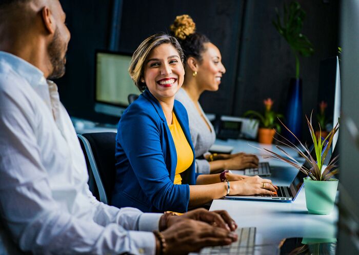 Three adults in a brightly lit office, one smiling, sharing a realization moment while working on laptops.