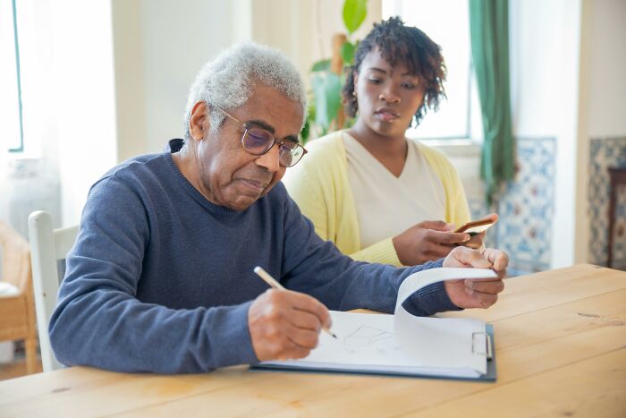 Elderly man writing notes while a woman looks on, both pondering adult realizations at a table.