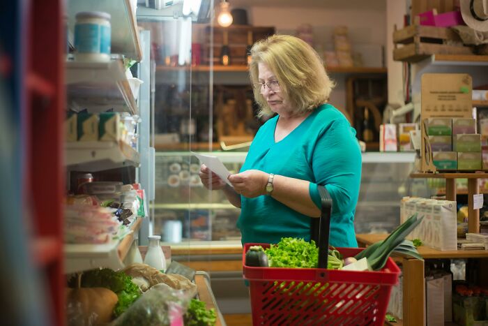 Woman shopping smartly in a grocery store, checking a list, surrounded by fresh vegetables, to slash her household budget.