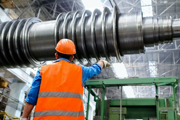Worker in an orange vest and hard hat, inspecting large industrial machinery in a factory setting.