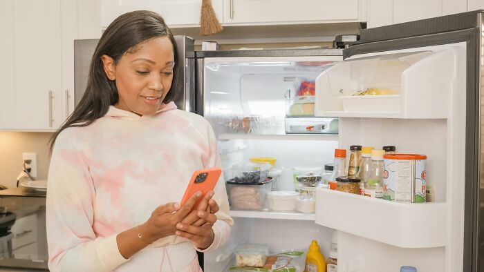 Woman using phone while organizing her fridge to slash household budget smartly.