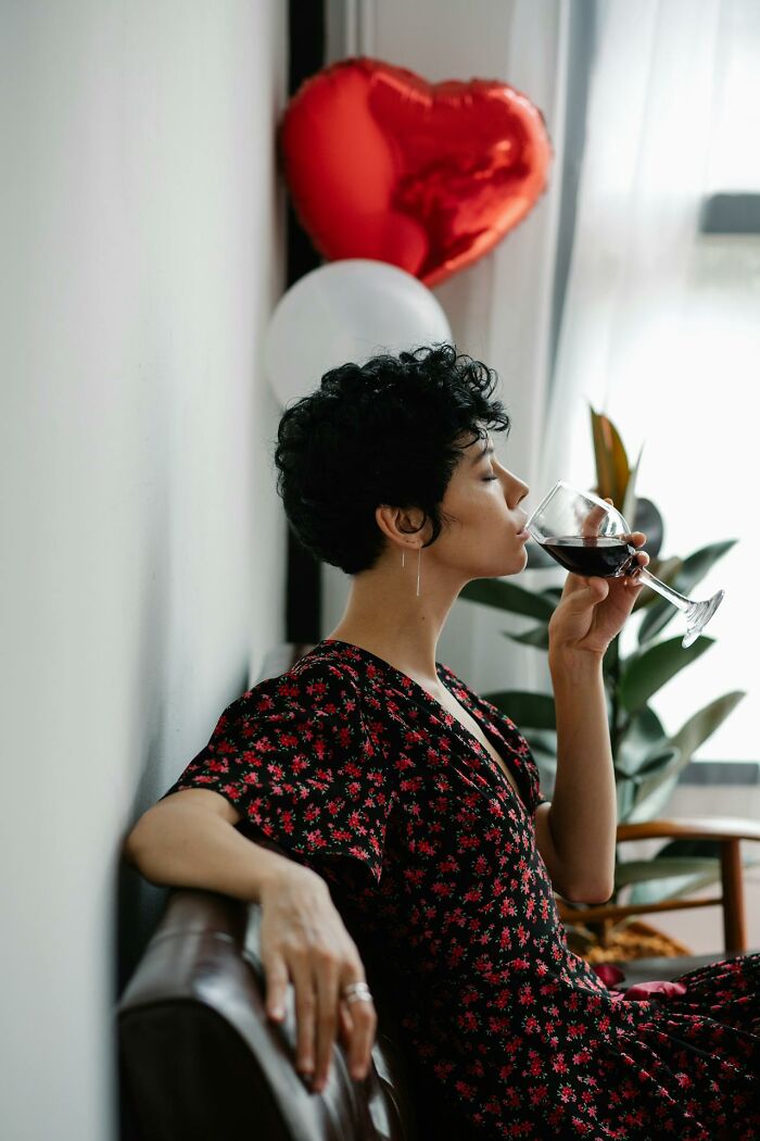 Person in a floral dress sitting on a couch, drinking red wine with heart-shaped balloons in the background.