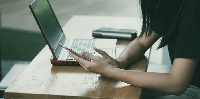 Person using a laptop and holding a smartphone at a wooden desk, illustrating technology use.