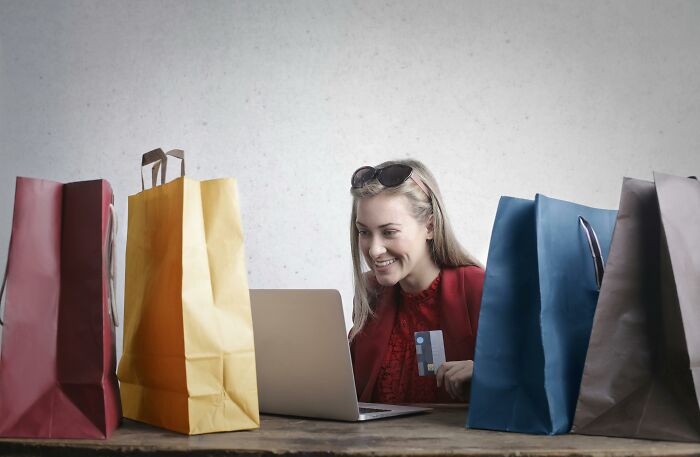 Woman shopping online at a laptop surrounded by colorful bags, discussing controversial conspiracy theories.