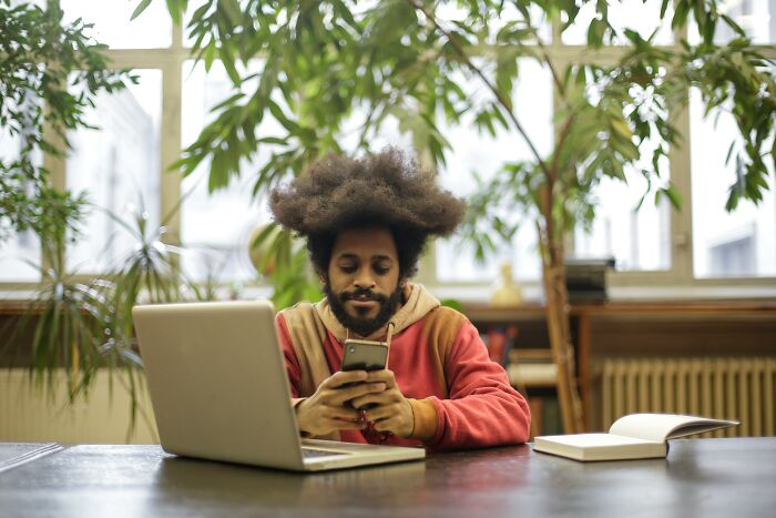 Man in a red hoodie using a smartphone at a table with a laptop, evoking 2000s teen nostalgia.
