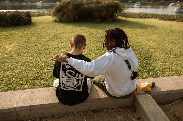 Two men sitting on a ledge by the water, one with an arm around the other, showing friendship and support.