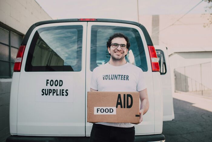 Volunteer holding a food aid box in front of a van labeled "Food Supplies," showcasing smart household budget strategies.