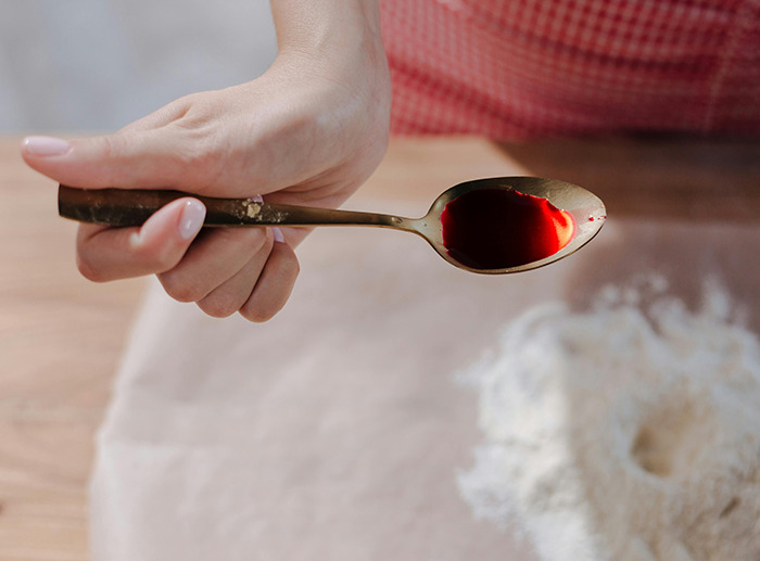 Hand holding a spoon with red dye No. 3 over a baking surface with flour.