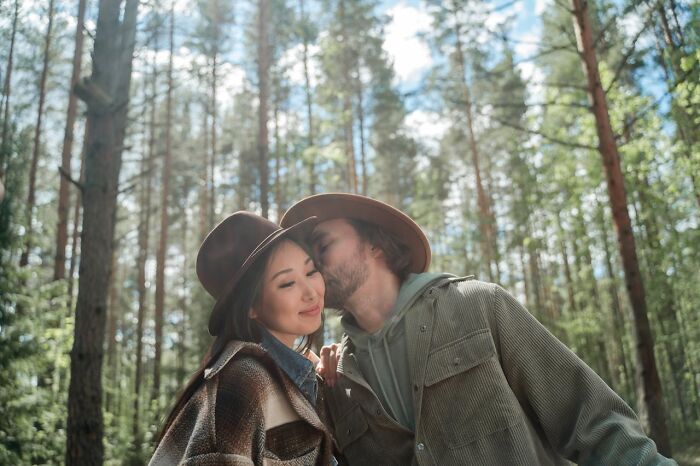 A man kisses a woman's cheek in a wooded area, both wearing hats, illustrating non-s****l acts that turn women on.