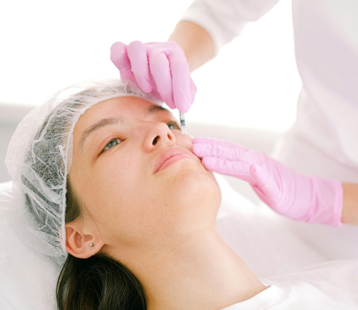 Woman receiving a face treatment, wearing a hair cap, with practitioner in pink gloves administering an injection.