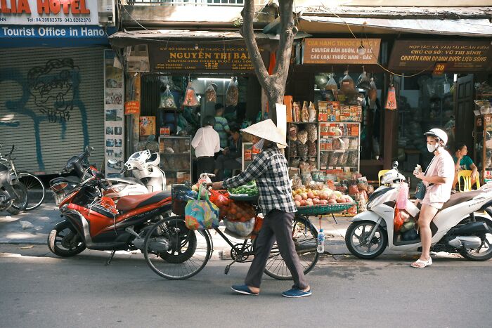 Street vendor in Vietnam with a fruit cart, passing by parked scooters and small shops.