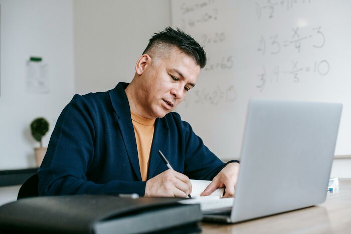 A teacher in a classroom writing notes in a notebook, with math equations on the board, sitting at a desk with a laptop.