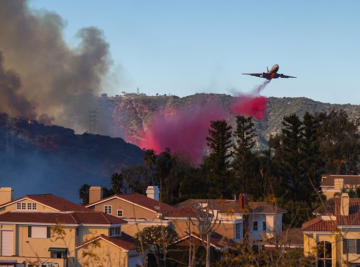 A firefighting plane drops fire retardant over hills near homes, combating wildfires and aiding private firefighters' efforts. A firefighting plane drops fire retardant over hills near homes, combating wildfires and aiding private firefighters' efforts.
