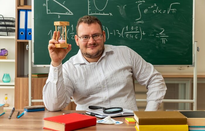 Teacher holding hourglass in classroom, seated in front of a chalkboard with math equations, surrounded by books and stationery.