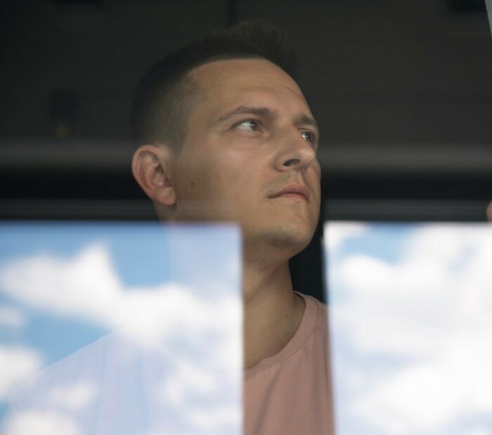 Man looking thoughtfully out a window against a cloudy sky, reflecting on secret mission in private yard.