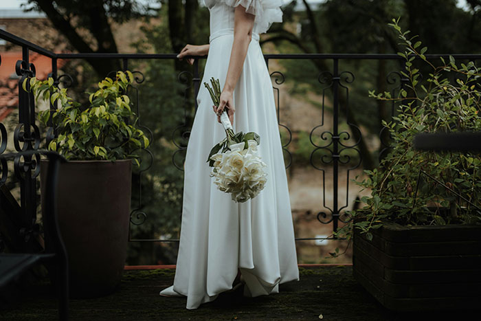 Bride in a white gown holding a bouquet, standing on a balcony with plants, reflecting on wedding decisions.