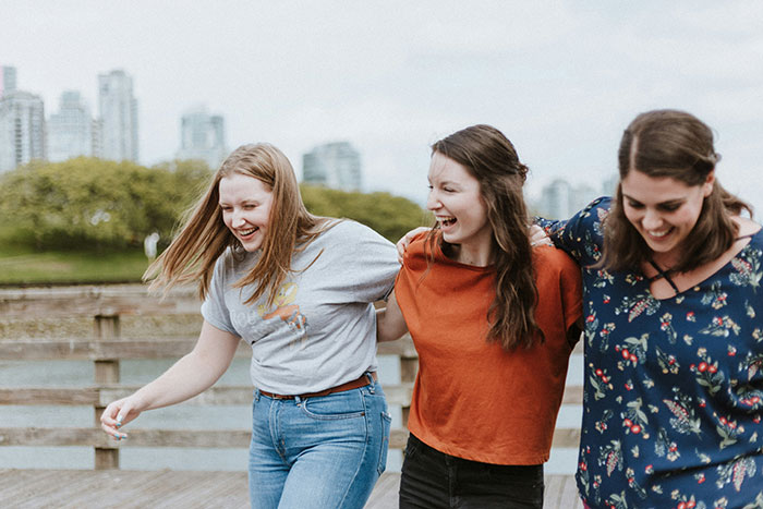 Three friends laughing and walking together on a wooden path with a city skyline in the background.