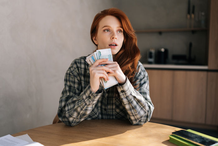 Person holding a stack of money thoughtfully at a kitchen table, related to reimbursement and expense receipts. Person holding a stack of money thoughtfully at a kitchen table, related to reimbursement and expense receipts.