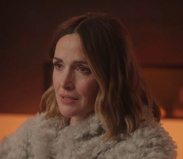 Rose Byrne in a fur coat, looking thoughtful in a warmly lit setting.