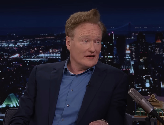 Conan O&rsquo;Brien discussing film at Sundance, wearing a blue shirt and blazer, city skyline in the background.