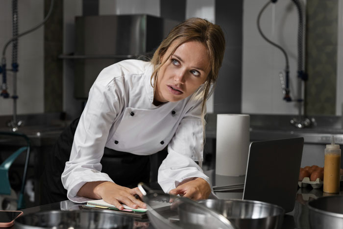 Wedding caterer in kitchen looking upset, wearing a chef's jacket, surrounded by cooking tools.