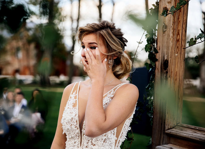 Bride in a white lace dress looking emotional outdoors, surrounded by greenery.