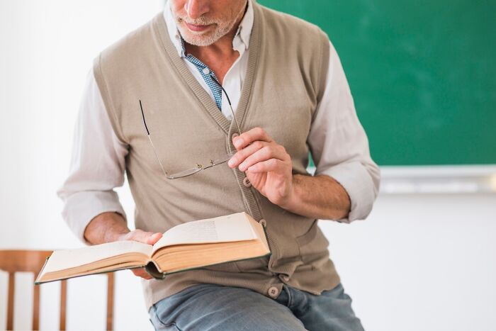 Teacher reading a book, holding glasses in a classroom, with a focus on crazy things teachers did in school.