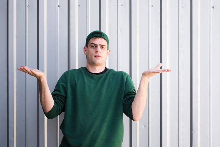 Man in green sweatshirt and cap shrugging in confusion, standing against a metal wall. Man in green sweatshirt and cap shrugging in confusion, standing against a metal wall.
