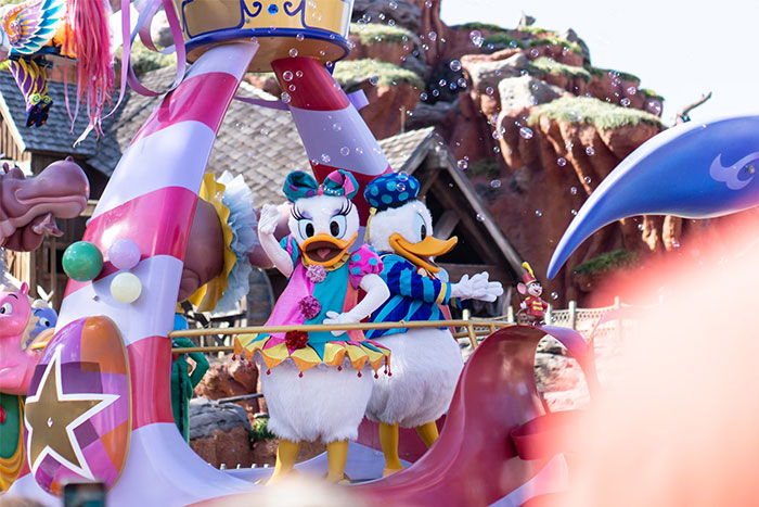 Donald Duck and Daisy Duck in colorful costumes smiling and waving during a theme park parade with bubbles floating around.