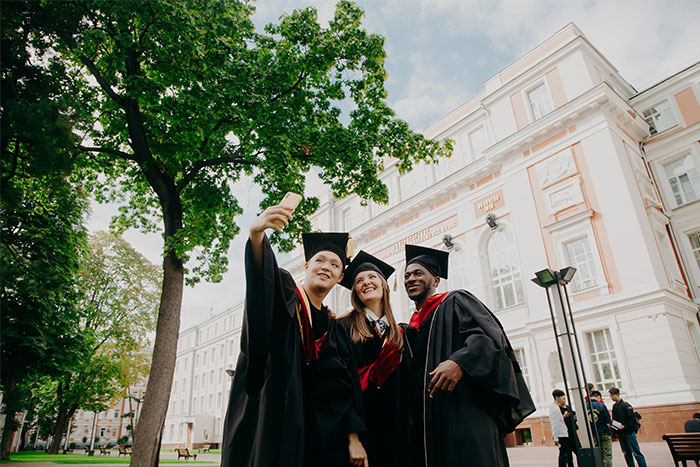 Graduates in caps and gowns taking timestamped pictures outside a historic university building on a sunny day.