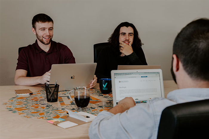 Three colleagues collaborate around a table with laptops, discussing timestamped pictures and creative ideas.