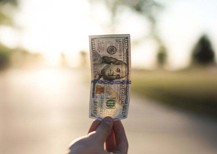 Hand holding a hundred-dollar bill outdoors with a blurred background, illustrating moments that saved people later.