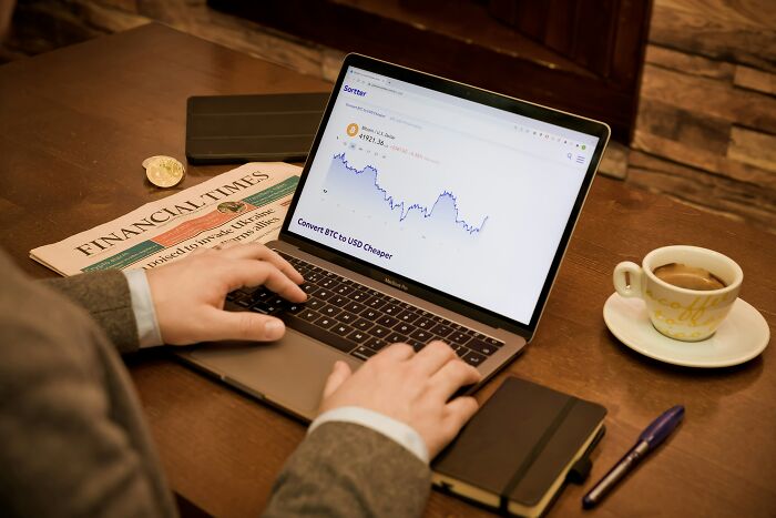 Person analyzing financial data on a laptop, with a Financial Times newspaper and coffee, reflecting financial stereotypes.