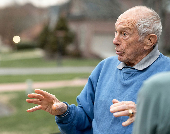 Elderly man in a blue sweater gesturing animatedly outdoors.