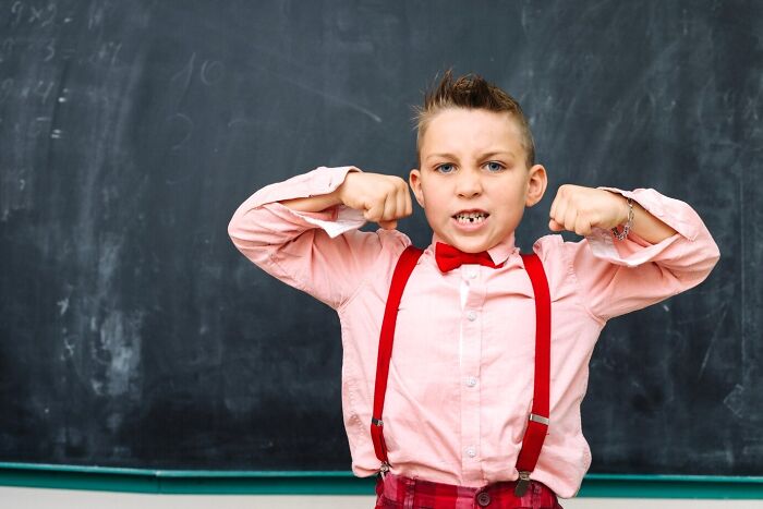 Child in a classroom flexing muscles, showcasing unique behavior in school, wearing a pink shirt and red suspenders.
