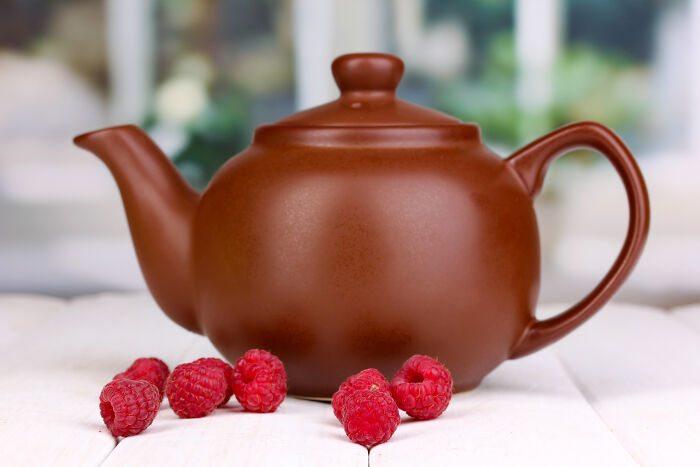 Brown teapot surrounded by fresh raspberries.