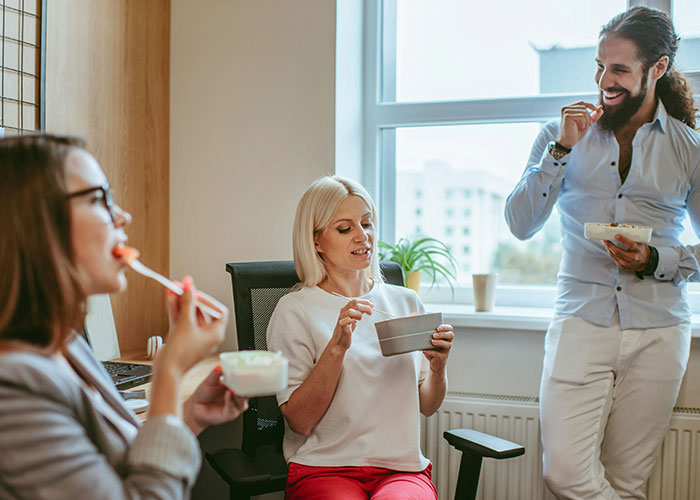 Office employees enjoying vegetarian snacks together, sitting and standing by a window.