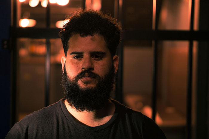 Man with a beard standing in dim light against a barred background, conveying dark secrets atmosphere.