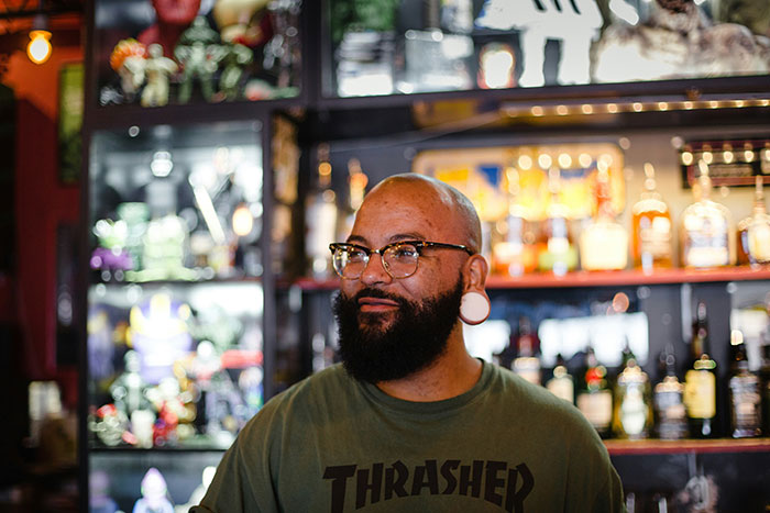 Man in glasses and green shirt standing at a bar, surrounded by bottles and colorful decorations, smiling casually.