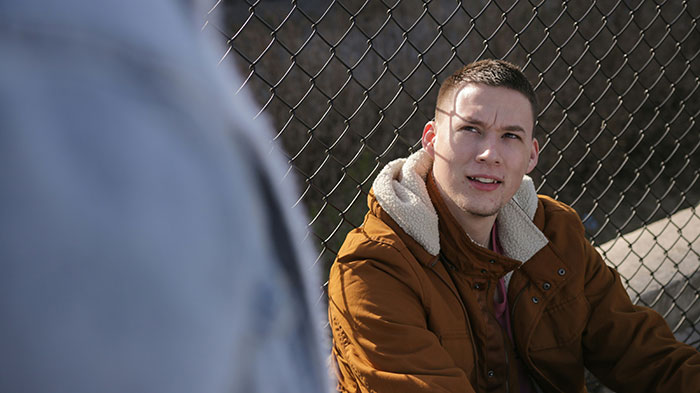 Man in a brown jacket sitting by a chain-link fence, discussing dark secrets confessed while drunk.