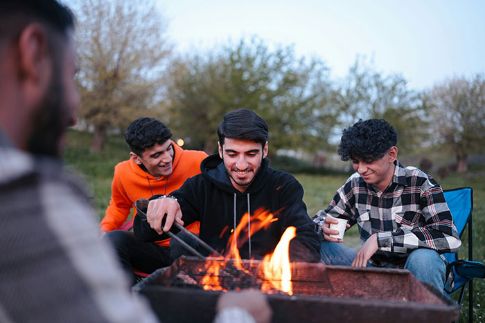 Three young men sitting around a campfire, enjoying a drink, and sharing dark secrets in a relaxed outdoor setting.