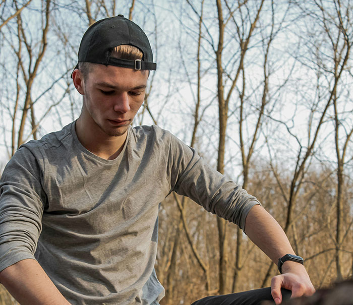 Young man in a cap and gray shirt sitting outdoors, looking thoughtful amidst bare trees.