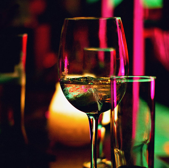 Wine glass on a dimly lit bar counter, reflecting colorful lights, symbolizing dark secrets and confessions.