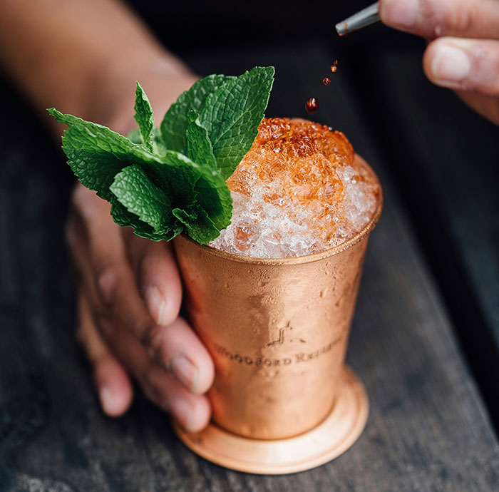 Copper cup with a cocktail, fresh mint garnish, and someone adding bitters, illustrating dark secrets theme.